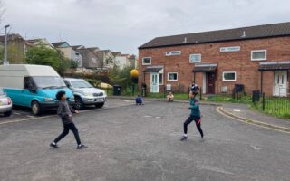 Two older boys playing football in a cup-de-sac with other children, houses and parked vehicles in the background
