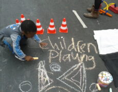 A small child is chalking on a street, the words Children Playing are written on the street surface and behind the child are some mini toy traffic cones