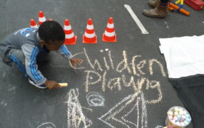 A small child is chalking on a street, the words Children Playing are written on the street surface and behind the child are some mini toy traffic cones