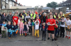 A group of people of various ages including children are looking at the camera happily. They are outside and the words Play Street are written in large chalk letters on a wall behind them.